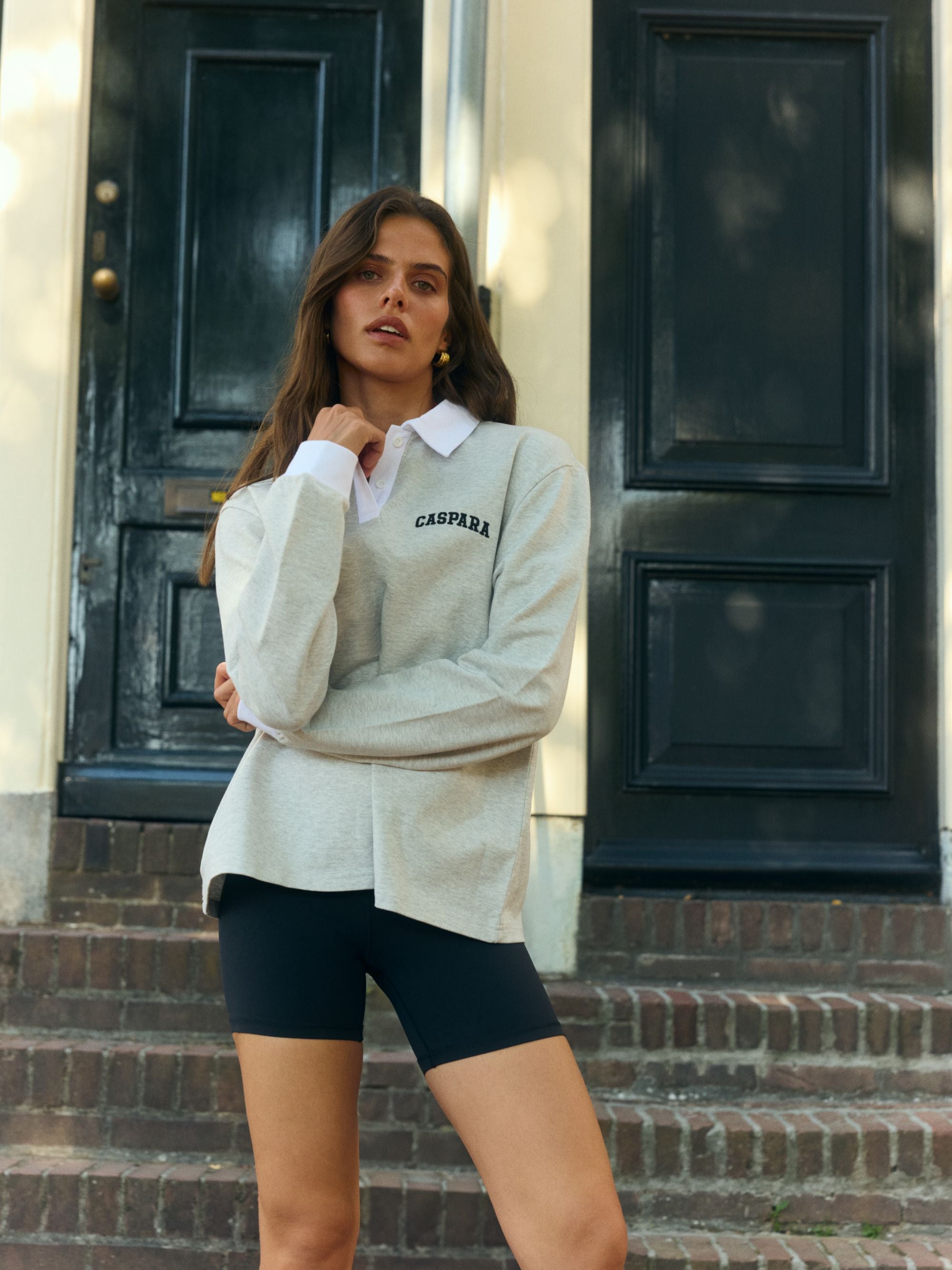 Woman wearing a grey CASPARA logo rugby shirt with a white collar and black biker shorts outdoors.